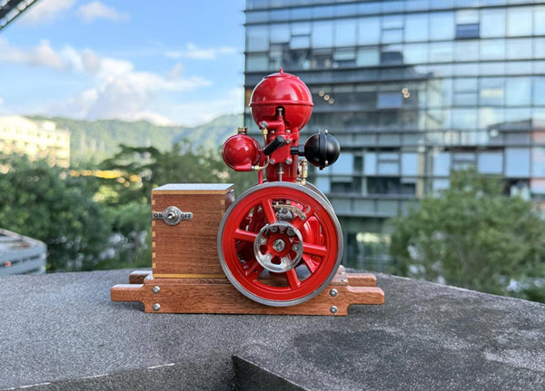 Red model steam engine with flywheel and wooden base, displayed outdoors