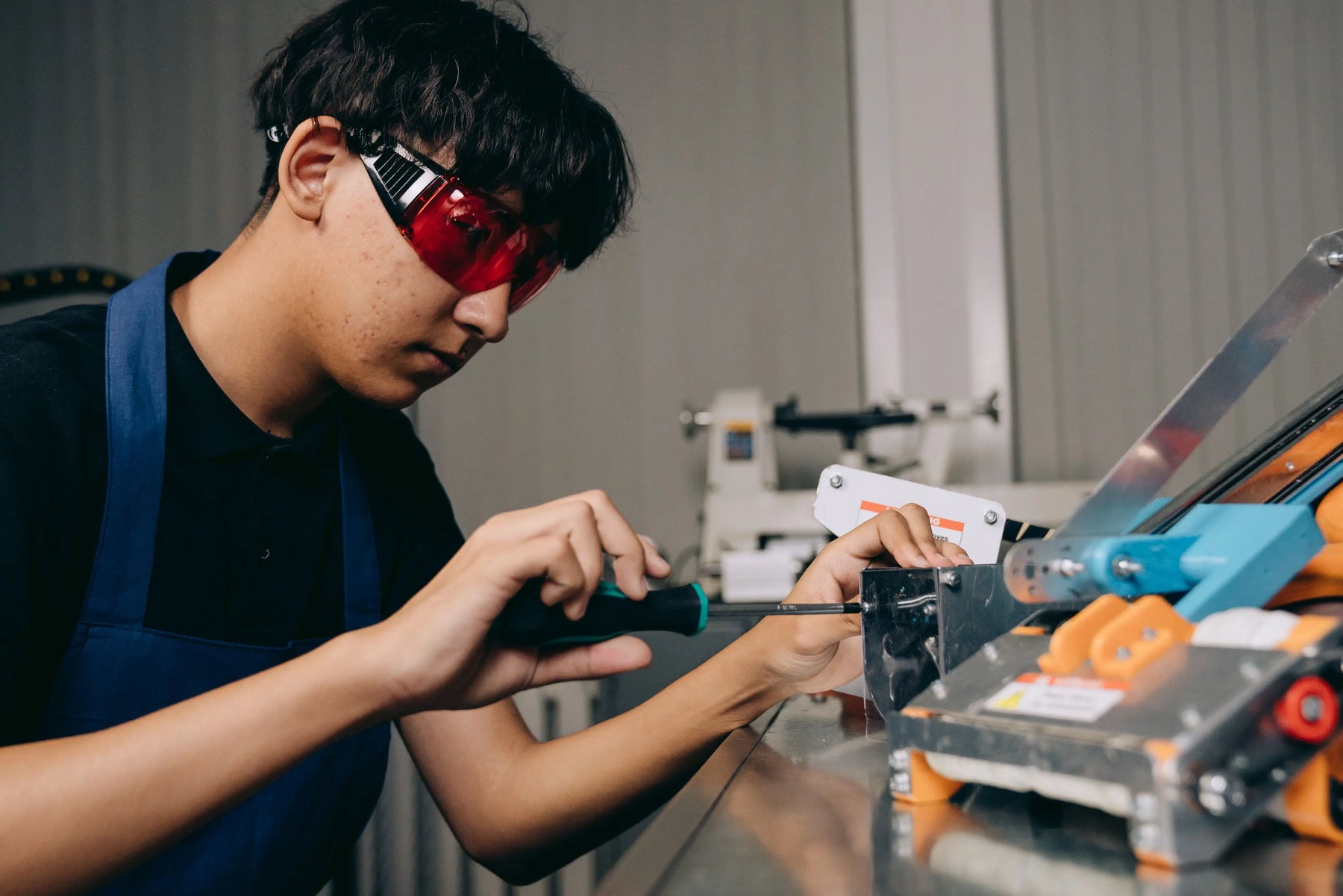 A person wearing red safety glasses uses a screwdriver to adjust a metal mechanism at a workshop bench.