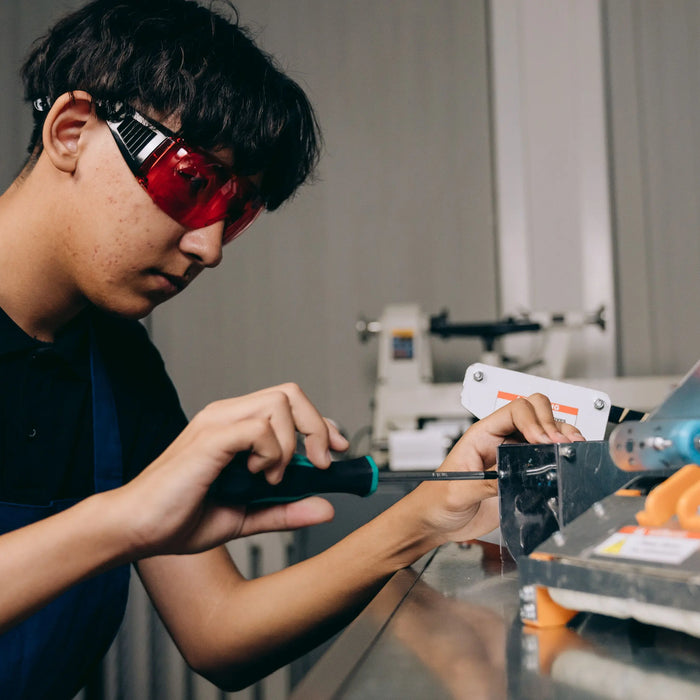 A person wearing red safety glasses uses a screwdriver to adjust a metal mechanism at a workshop bench.