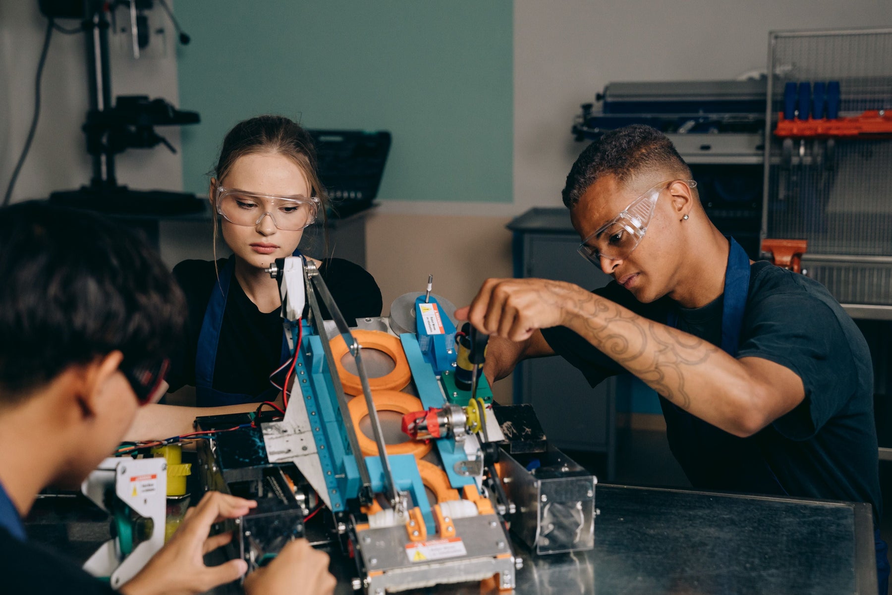 Three young people wearing safety glasses work together on a mechanical project at a workshop table.