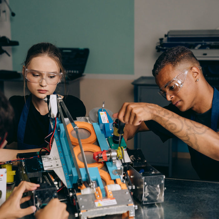 Three young people wearing safety glasses work together on a mechanical project at a workshop table.